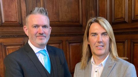 Two men stand in front of a panelled wall. The man on the left is wearing a black suit with a blue tie with short grey hair, and the man on the right is wearing a grey suit and has long blonde hair