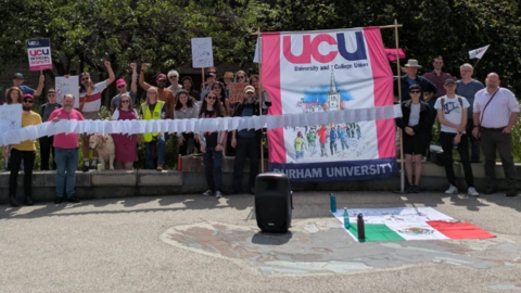 UCU members forming a picket line outside Durham University. Several people in the large group are carrying placards, while two people are supporting a large pink and white UCU banner.