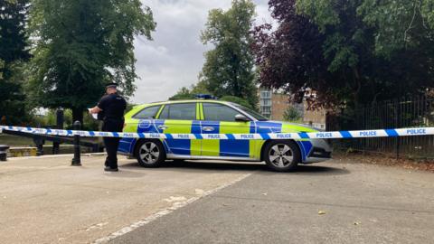Police tape in the foreground with a police car beyond it, blocking a road. A police officer is walking behind the car looking at some paper. In the background are trees and a building.