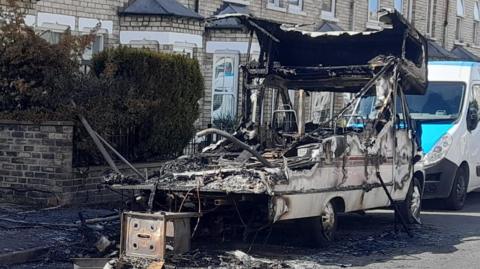 The image shows the aftermath of a vehicle fire on a residential street outside a row of terraced houses. The street is lined with parked vehicles. In the centre-right of the image is a severely burnt-out campervan. The vehicle is destroyed, with the metal frame exposed and blackened.