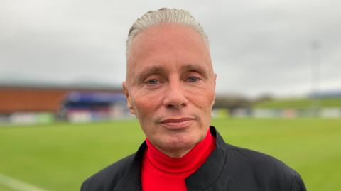 Barrie Drewitt-Barlow has blond, slicked back hair. He is wearing a black suit jacket over a red roll-neck top. He looks serious while standing in front of the pitch at Maldon and Tiptree Football Club.
