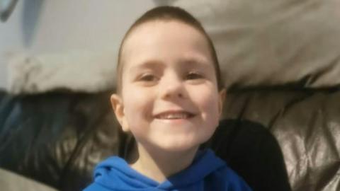 Smiling young boy with dark hair, wearing a blue top pictured on leather sofa