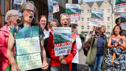 Protesters opposed to the development outside the Scottish Parliament. There are eight women and two men in a group, chanting and carrying placards reading Say No to Flamingo Land.
