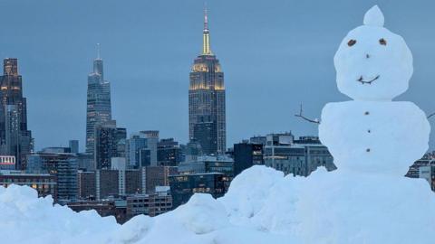 A close-up of a snowman in a park in Hoboken, New Jersey. The skyline of midtown Manhattan and the Empire State Building in New York City in the background. The lights in the buildings are on.