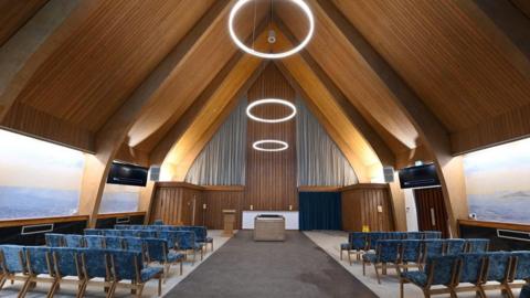 Interior of Halo Hall. A large modern hall with a vaulted wooden ceiling, large pictures on the walls and rows of blue velvet chairs.