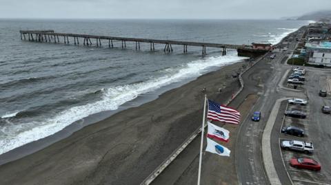 An aerial photo show an American flag on a pole with the ocean behind and a pier stretching over it on an overcast day