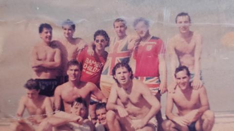 A faded photograph of a group of men on a beach in swimming trunks, while some of them wear England T-shirts.