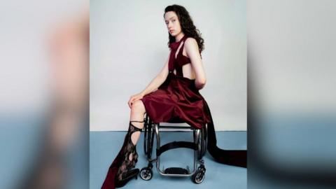 Alice Dyer has curly brown hair and is sitting sideways in her wheelchair. She's posing for a photo shoot wearing a dark red short dress with cutouts. She has on black lace socks