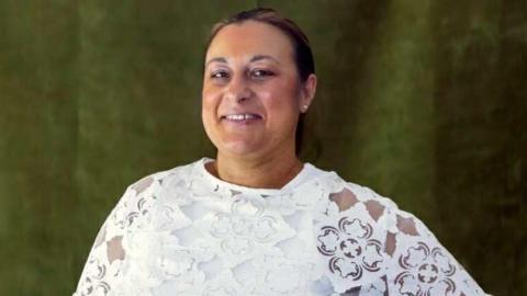 A woman with tied-back brown hair and wearing a white top, smiles while looking at the camera.