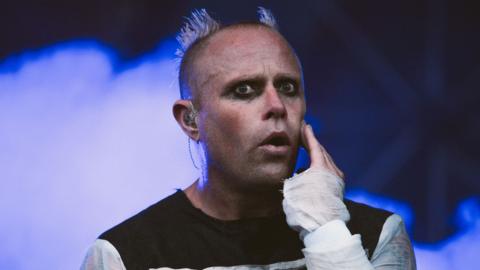 Keith Flint is on stage while wearing a black top with white sleeves. He has dark eyeliner on and his head is shaved, except for two parts which stand up like devil horns. Blue smoke billows behind him.