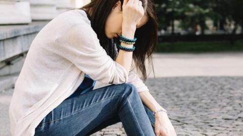 A stock image showing a girl with long dark hair. She is sitting on stone steps and leaning forward over her raised legs. Her right elbow is resting on her right knee so her hand shields her face. She is wearing a cream cardigan over a blue top and blue jeans