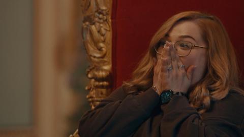 Sarah Taylor, a woman in her thirties, sits on a throne-like chair with her hands covering her face in disbelief.