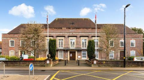 A large red brick building with an iron railing balcony. There are flag poles on either side of the entrance. 