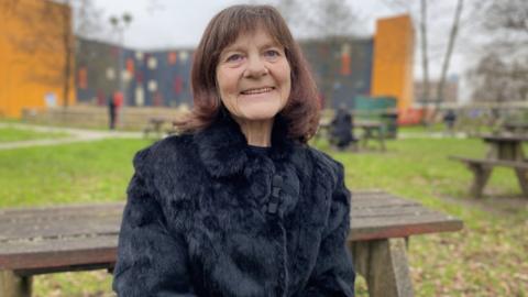 Hazel, 65, has a brown bobbed haircut and is wearing a black fluffy coat. She is sitting on a picnic bench in the grounds of Cardiff's University Hospital of Wales.  