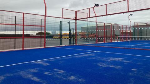 A blue floored padel court, surrounded by red fences. 