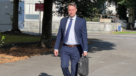 Huw Orphan, wearing a blue suit, white shirt and tie and carrying a bag, walks near Cardiff Crown Court and looks at the camera