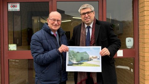 Two men standing outside a set of double doors hold up an poster showing the design of a proposed new sports facility.