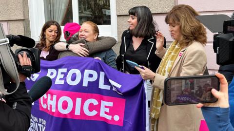 Five women celebrate with hugs next to a banner saying 'people, choice' with news cameras around them, outside on the street in Jersey on Thursday.