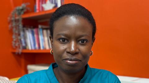 Faty Kane looks a the camera. She has very short black hair and is wearing a blue shirt, sitting in front of an orange wall.