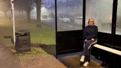 Councillor Vicky Oakley sitting at a bus stop in Pegswood. She is wearing a dark top and tracksuit bottoms and looks unhappy. It is getting dark and the night seems quite foggy.