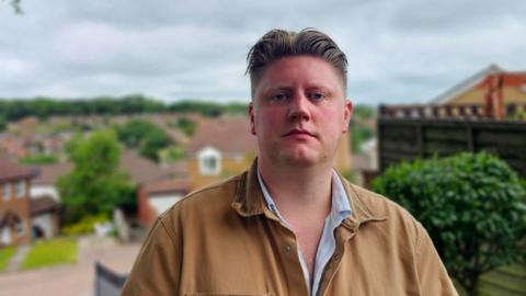 Councillor Robbie Lammas stands with his council ward in the distance behind him. In the foreground Councillor Lammas is wearing a beige jacket and looking towards the camera 