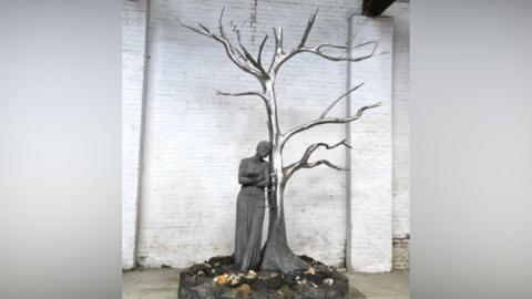 A statue including a shiny metal tree without leaves and a person leaning up against it on a scenic base with a white brick wall behind it