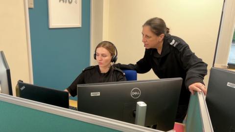 TVP PC Harley Edwards and Sgt Dom Lucas from the RVR team in front of a computer. PC Edwards has a headset on and appears to be on a call. Sgt Lucal is over her shoulder.