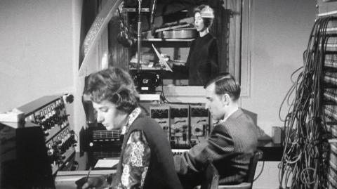 Three members of staff working in the Radiophonic workshop. Two are seated at a desk and one, at the back, is in a recording booth.