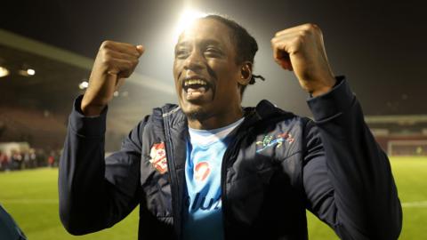 Nile Ranger, who has short black, hair and is wearing a blue football shirt and a black raincoat, raises both fists in a triumph at the side of the pitch. The pitch and a stand can be seen behind him.