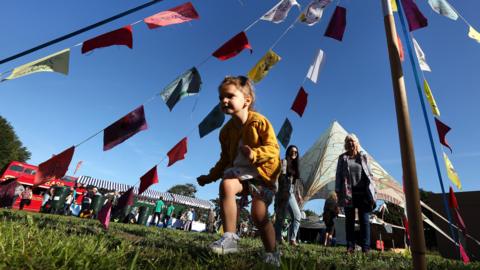 A young girl is dressed in a yellow cardigan and is running under a maypole with brightly coloured bunting. Stalls can be seen in the background with people perusing them.