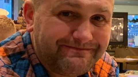 A close-up of a man's face. He is smiling at the camera, wearing a blue and orange checked shirt.