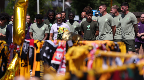 A number of men in football training tops can be seen across the top of the picture, looking solemn.
The bottom half of the picture is partially out of focus, but is centred on football scarves draped over yellow barriers