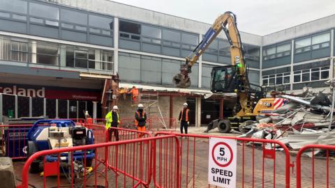 Photograph of demolition work at the former Peacocks store at Wythenshawe Civic Centre. The image shows rubble, a digger, fencing and construction workers in hard hats. 