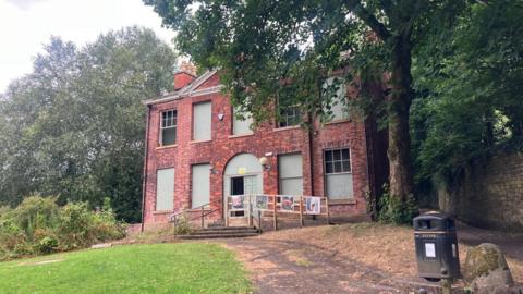 Photograph of Rock Hall in the Moses Gate Country Park in Farnworth. The image shows boarded-up windows and the building is surrounded by trees.