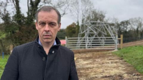 A man wearing a black coat is looking at the camera. Behind him is a half constructed electricity pylon