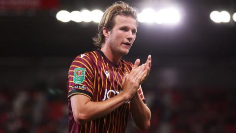 Tommy Leigh has his mouth open a little and claps his hands together in front of his chest. He has a band holding his hair off his face and wears a claret and amber football shirt