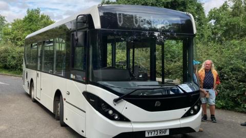 A white and black single decker bus next to green bushes with two mean standing next to the passenger door