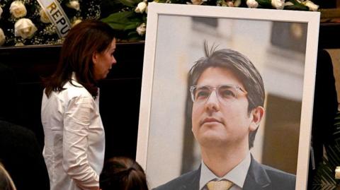A woman in a white jacket stares at a large photograph of deceased senator Miguel Uribe, which has been put up on a stand at his funeral. 