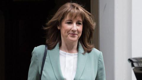 Rachel Reeves walks out of Number 10 Downing Street wearing a white blouse and light blue jacket smiling slightly with her mouth closed