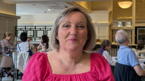 Tracy Tomlinson with a grey coloured bob haircut wearing a fuscia pink top. She is sitting in a busy restaurant and is smiling.