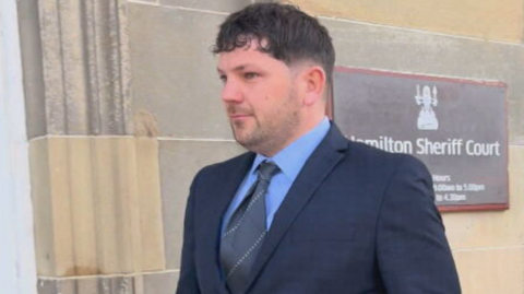 Tom Nicholson Jr is wearing a navy suit, light blue shirt and blue tie. He is leaving Hamilton Sheriff Court. He is pictured side on and is looking straight ahead. 