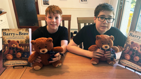 Oliver and Daniel Martin are sitting at a table holding brown teddy bears with scarves on. Each has a Barry the Brave book beside them featuring the same bear on the front cover. Oliver has short, blond hair and Daniel has short, dark hair an black-rimmed glasses. They are both wearing black T-shirts.
