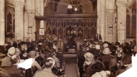 Black and white photo of a large audience in the church pews and a choir in the choir stalls with Reverend Ridge conducting them.