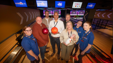 A group of people are standing on a lane in a bowling alley. Two of the men are holding bowling balls and a woman is holding a skittle.