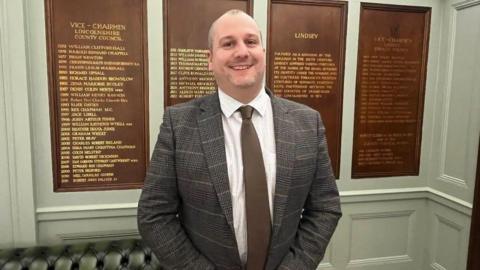 Councillor Carl Macey stands in a wood panelled room. He is wearing a grey checked suit and a brown tie. He is looking straight into the camera and is smiling.