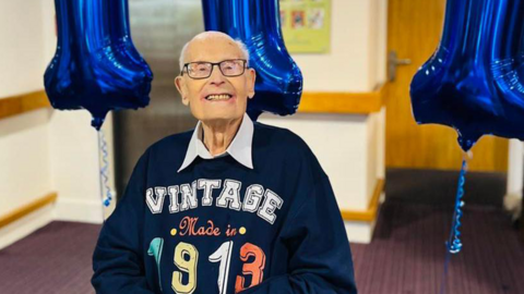 John Farrington smiling at the camera in a blue "vintage made in 1913" jumper, with three cakes spelling out '111' in front of him and blue balloons behind him