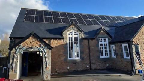 The roof of the parish hall in St Martin covered in solar panels as a man works up a ladder nearby. It is a stone building with large, white-framed windows and an arched entrance with a decorative framework.