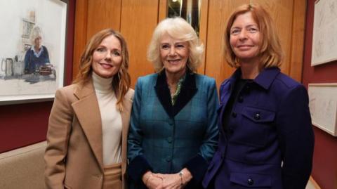 Geri Halliwell-Horner, the Queen and Maggie's chief executive Dame Laura Lee stand in a wood panelled booth. Horner-Halliwell wears a beige suit with a white turtle neck, the Queen wears a blue tweed suit with black colour and cuffs, Dame Laura is wearing a purple jacket and dark trousers. 