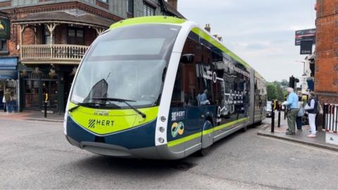 A Hert "trackless tram" driving along a road in St Albans. It is shaped like a tram with a pointed nose, but is effectively a two-carriage single-decker bus on wheels. It has lime green and navy blue livery, and has a a map of the locations where it stops on the side. People are waiting at a crossing to cross over the road. In the background are some old-fashioned looking shops.