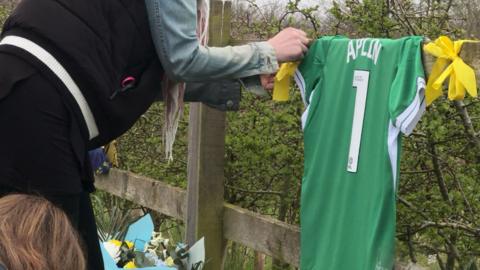 A green shirt with Amelia's name and the number 1 hangs from a fence whilst a woman signs it.